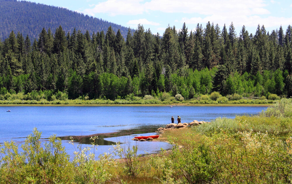 Two people are fishing in Nevada on the edge of a lake, near a red canoe. The lake is surrounded by tall green trees.