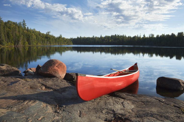 A red canoe on a rocky shore of a calm blue lake surrounded by trees under a wispy cloud-filled sky