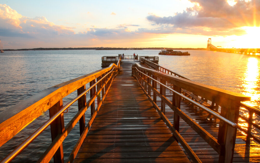 The sun rises and turns the water by a wooden pier golden. Several people stand, fishing, at the edge of the pier.