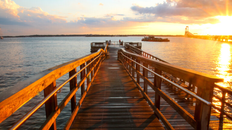 The sun rises and turns the water by a wooden pier golden. Several people stand, fishing, at the edge of the pier.