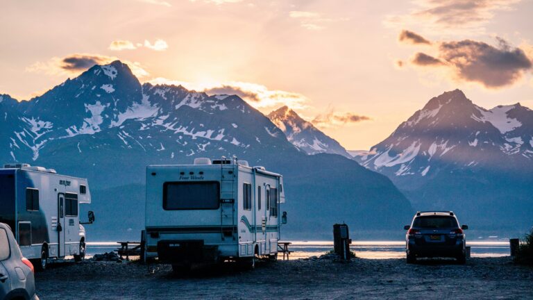 Two Class C motorhomes and two passenger cars are parked by the water in front of Mount Alice with the sun on some clouds.