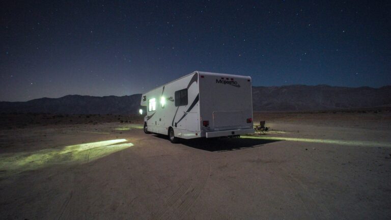 A white Class C motorhome with light streaming from its windows sits under a starry sky somewhere in the California desert.