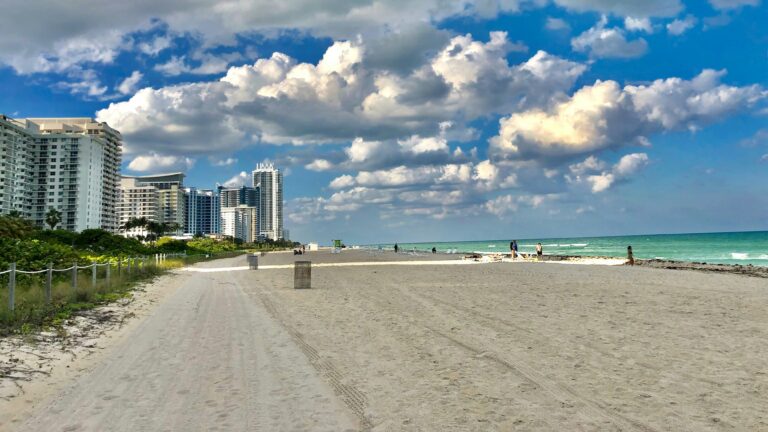 Tower resorts and hotels with the tide coming in toward a white sand beach covered in lounge chairs and lush palm trees.