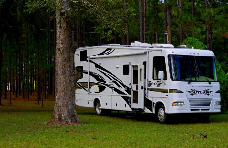 A white Class A Outlaw RV sits parked beneath tall pines with green grass growing all around.