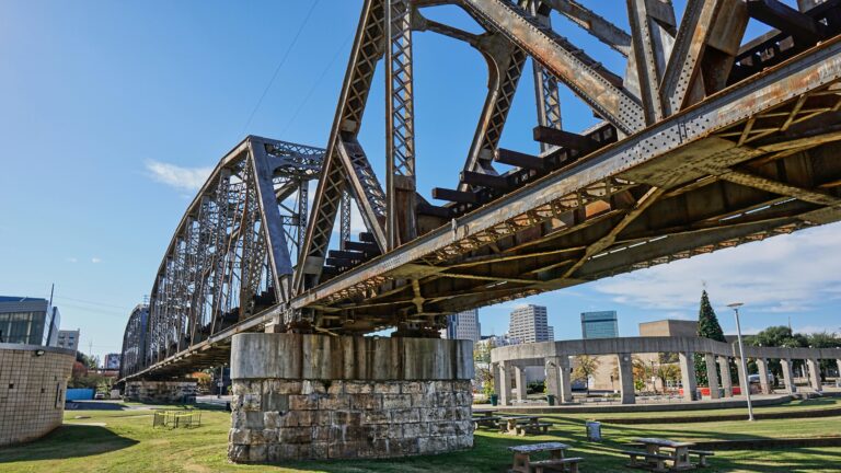 Metal suspension bridge with arches above green grass with the city skyline in the background.