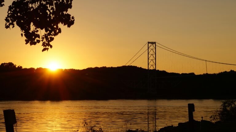 A river bank with the river flowing in the middle and a forest and a suspension bridge in the background at sunset.