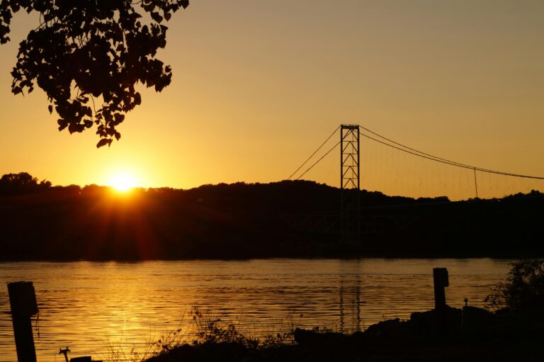 A river bank with the river flowing in the middle and a forest and a suspension bridge in the background at sunset.