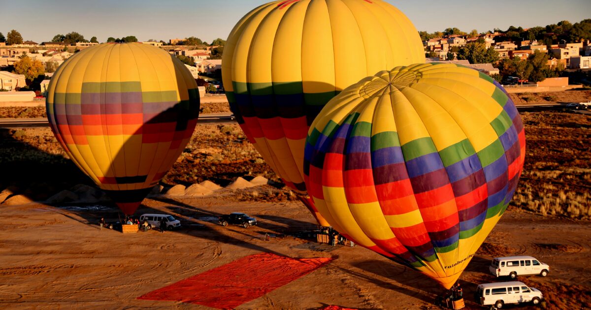 Three colorful hot air balloons on desert ground at sunrise, with red tarps and white vans nearby
