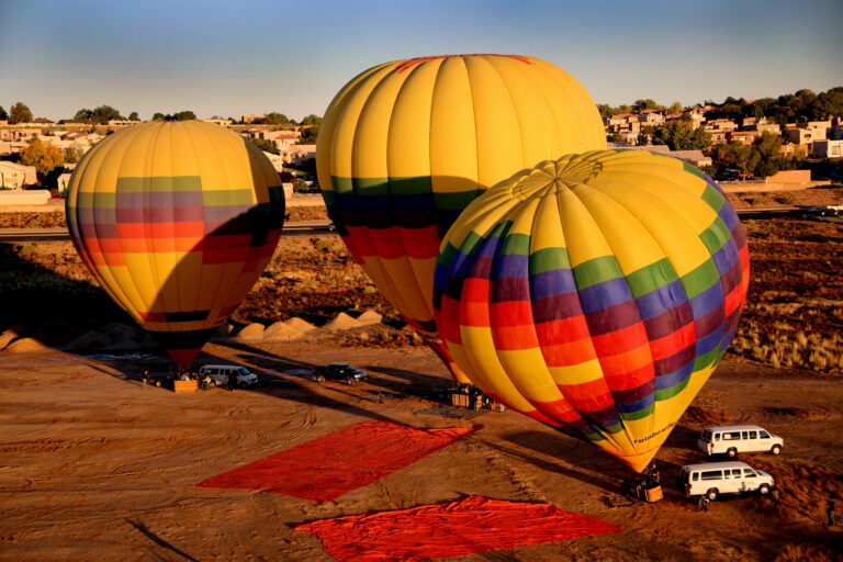 Three colorful hot air balloons on desert ground at sunrise, with red tarps and white vans nearby