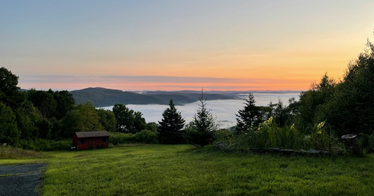 A red barn sits on a green expanse of grass with hills and fog in the background.