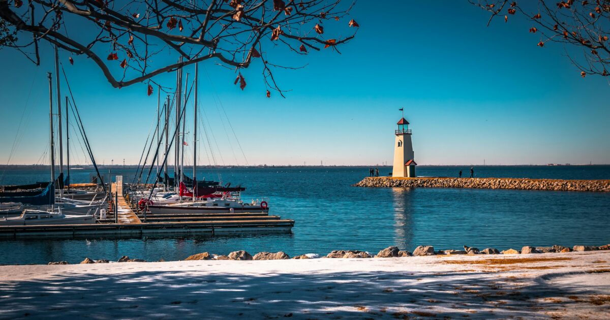 A shoreline next to a body of blue water, boats, and a lighthouse.