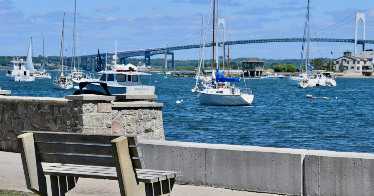 Sail and power boats sit at anchor with a bridge in the background under a radiant sky with a few clouds.