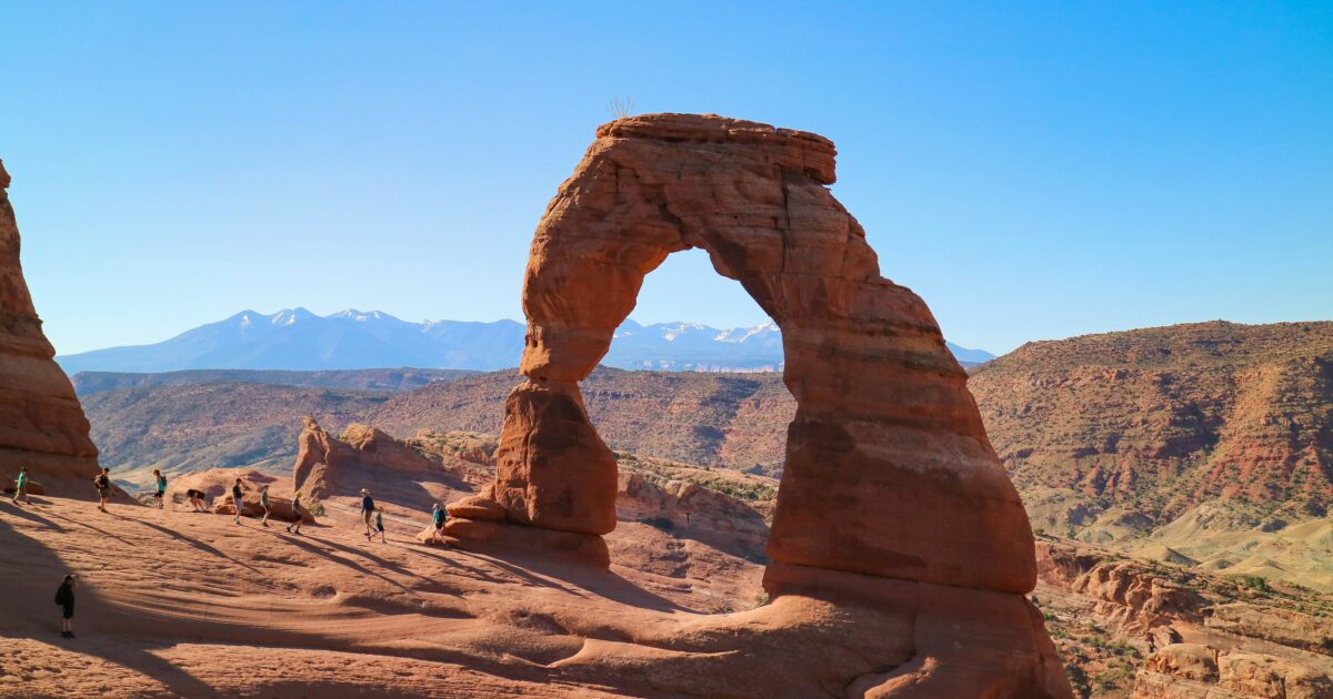 Red stone arch curves through a blue sky with mountains in the distance.