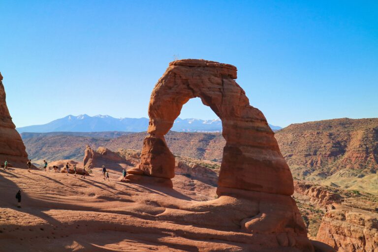 Red stone arch curves through a blue sky with mountains in the distance.