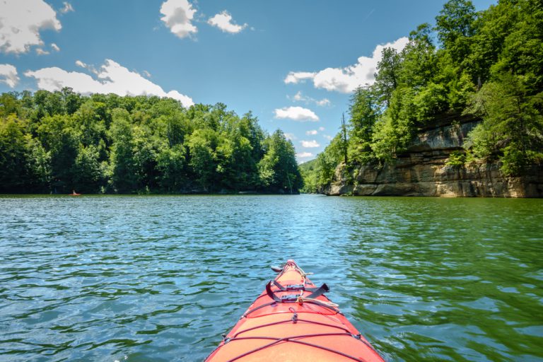 The front of a red kayak floating in a lake surrounded by tall green trees