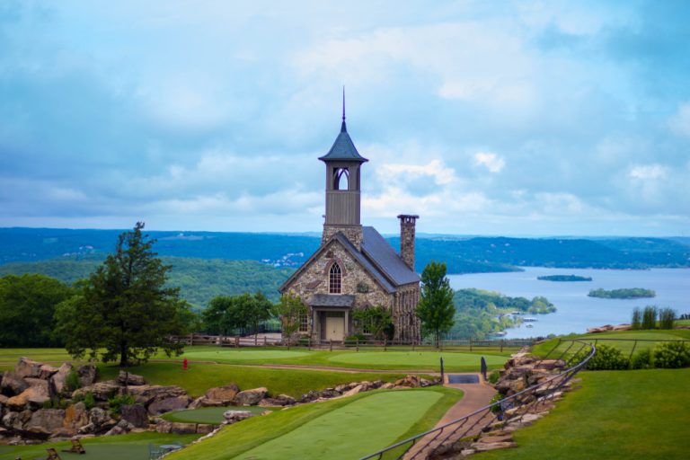 A stone building with a steeple sits on a grass covered, rocky hill overlooking a river in the distance.