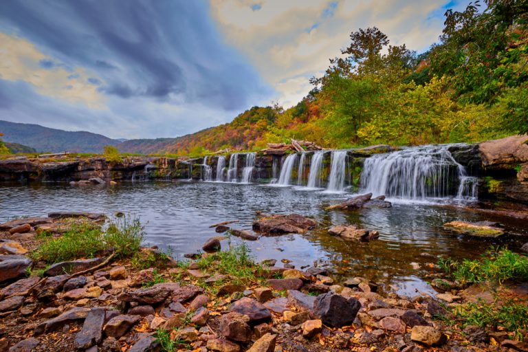 Water falls over a short rocky cliff. Boulders and rocks sit in the water below. Tall green and autumn colored trees stand on top of the cliff. Mountains can be seen in the distance.