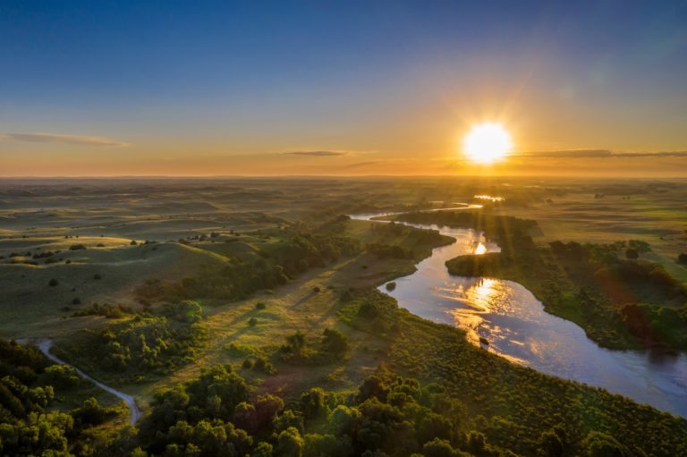 The sun rises over grassy hills with trees. A river meanders across the landscape.