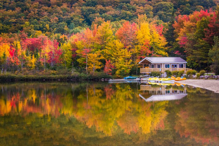 A boathouse on the edge of a lake that reflects the surrounding red, orange, and yellow colors of the trees