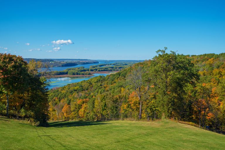 Green, yellow and orange trees on a rolling hill under a blue sky. A river flows in the distance between islands with trees.