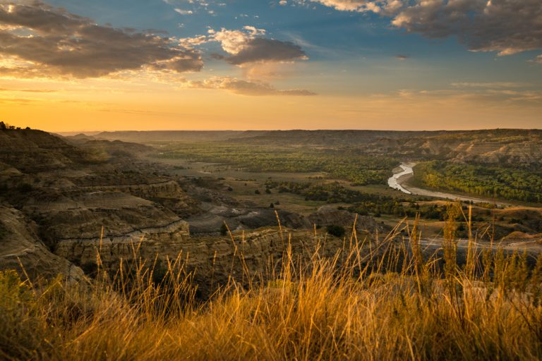 The sun rising over a rocky mountain. In the distance, a river flows surrounded by trees. Tall wheat-colored grasses sit in the foreground.