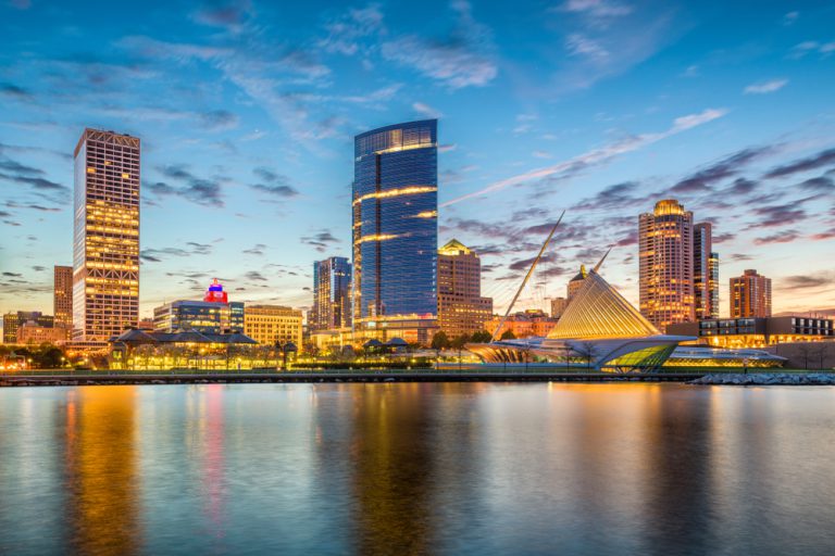 A lake reflects a cityscape at twilight under a blue sky dotted with gray clouds.