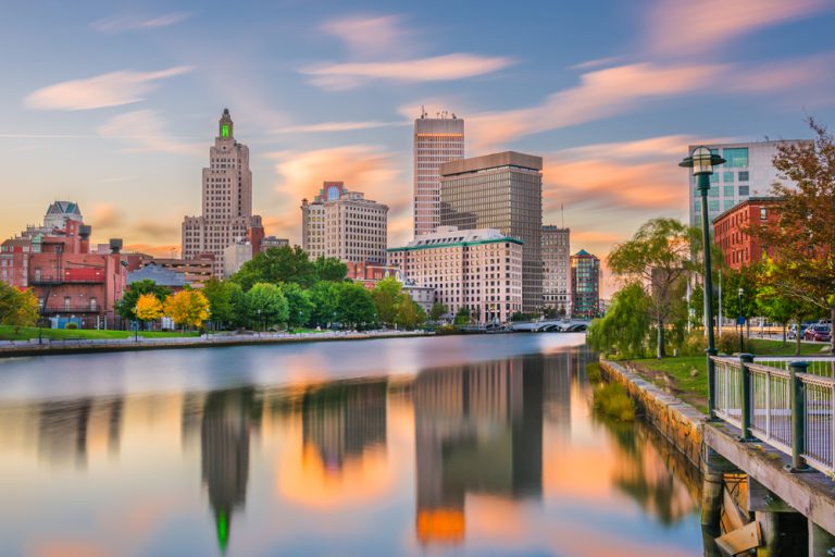 A river reflects a tall cityscape under pink clouds and a blue sky.