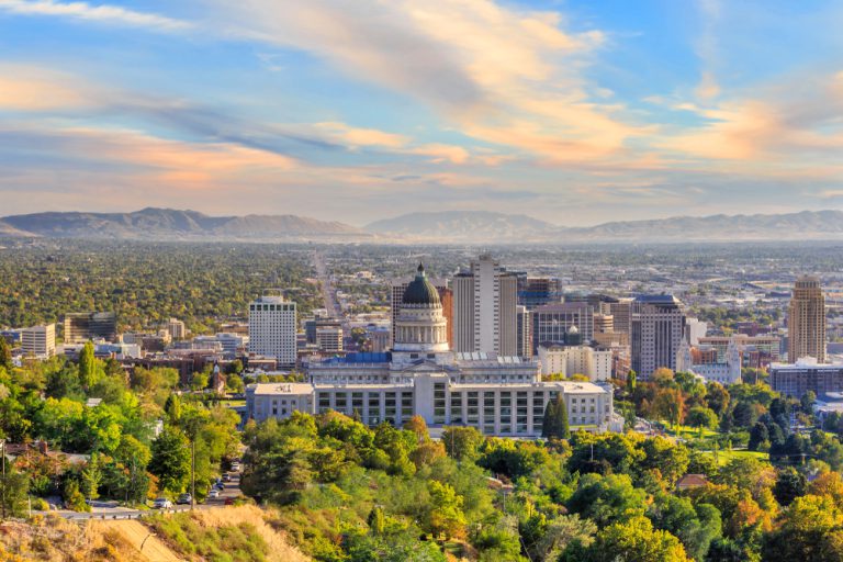 A dome-topped building sits behind green trees with a cityscape and mountains in the distance under a cloudy blue and golden sky.