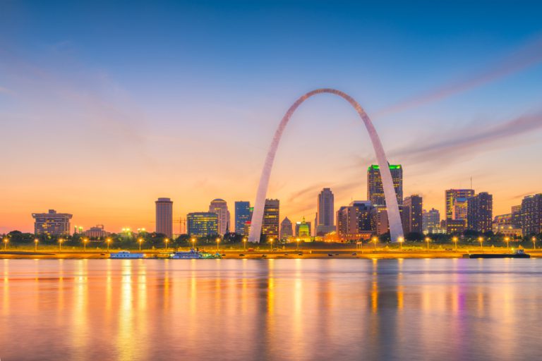 A downtown cityscape with a tall arch structure beside a river at dusk.