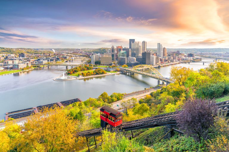 A red structure on a bridge surrounded by green trees overlooks a cityscape across the river.