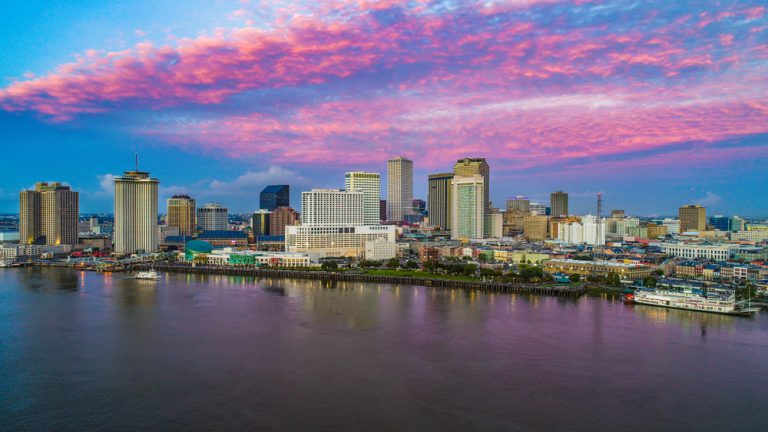 Cityscape on the shore of a wide river. A steamboat floats near the shoreline under pink clouds in a blue sky