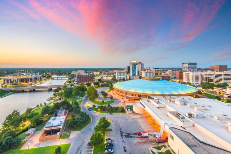 A circular building with a teal roof stands in a city beside a river under a purple and blue sky with pink clouds.