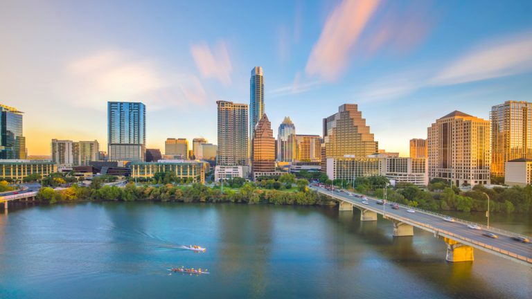 Kayakers float on the river toward a bridge that leads to a city under pink clouds and a blue sky.