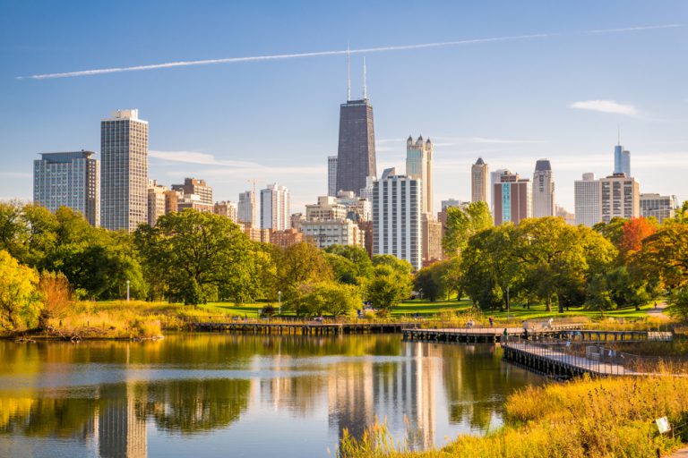 A pond surrounded by grass and autumn colored trees in front of a cityscape under a blue sky with wispy white clouds