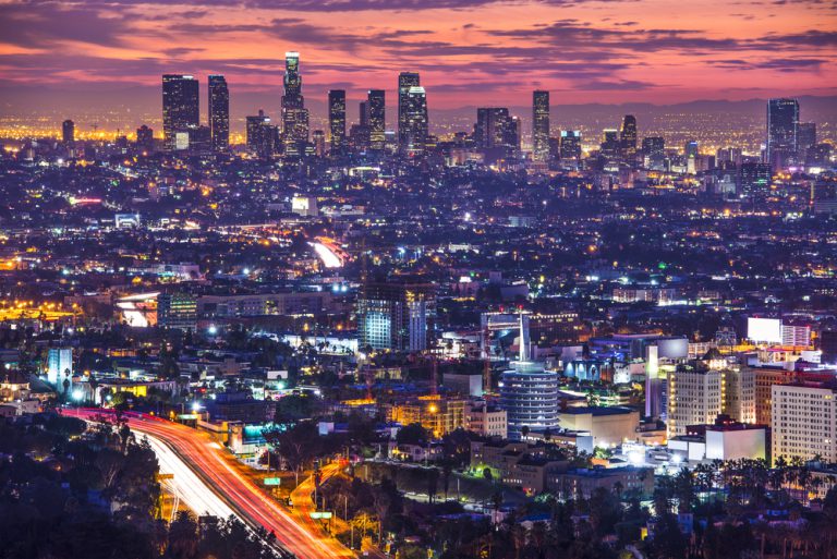 Brightly lit skyscrapers sparkle amid a sea of city lights with wispy clouds in the sky against the reddish glow of dawn.