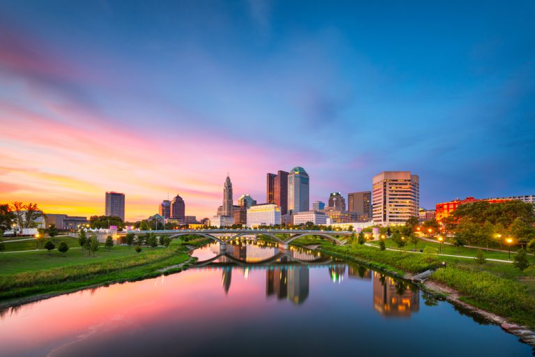 A river reflects a cityscape under a pink, purple and blue sky.