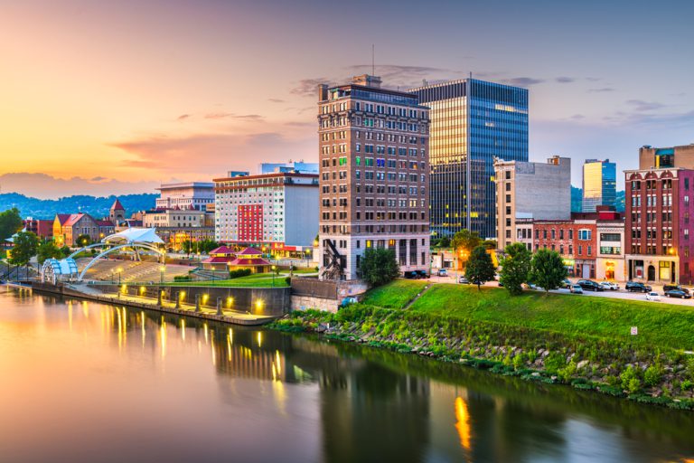 A glowing river at dusk flows along a bank of tall, colorful office buildings with a line of greenery in the foreground