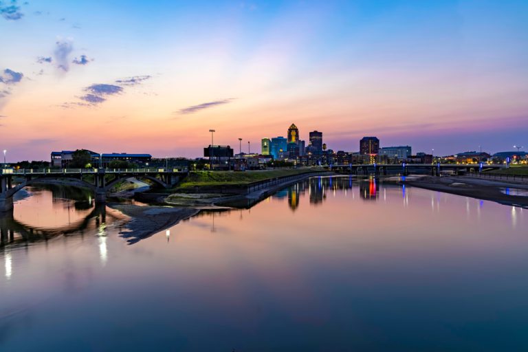 A bridge spans a calm, reflective body of water with a cityscape in the background under an orange and purple sunset.