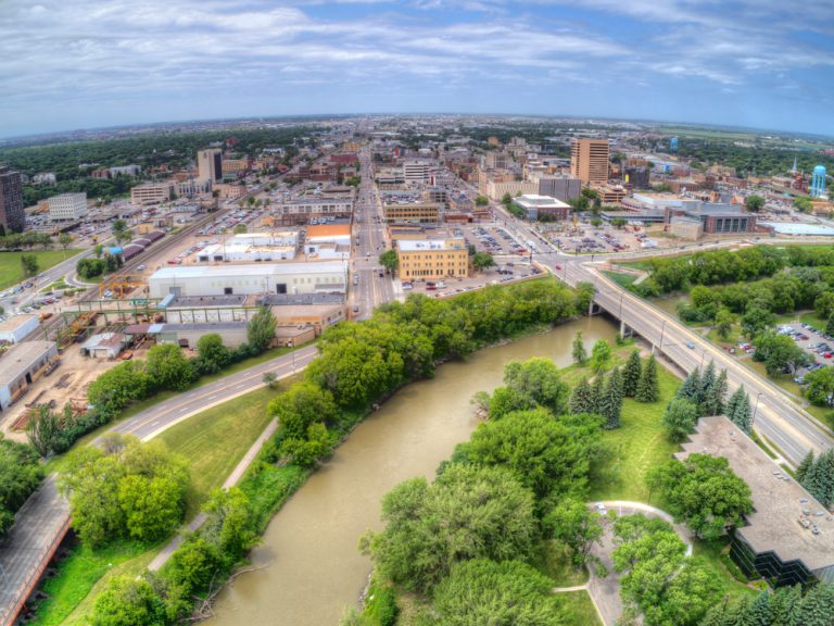 A river bordered by green trees runs beside a city under a blue sky with wispy white clouds.