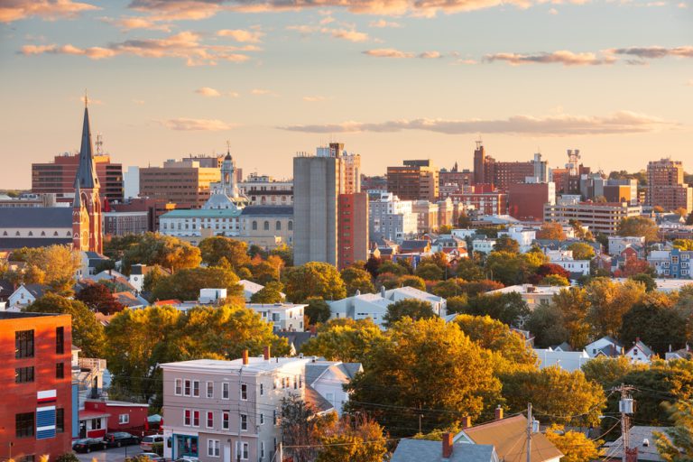 A city with homes, tall brick buildings, and green trees sits under a partly cloudy orange and blue sky at dusk.