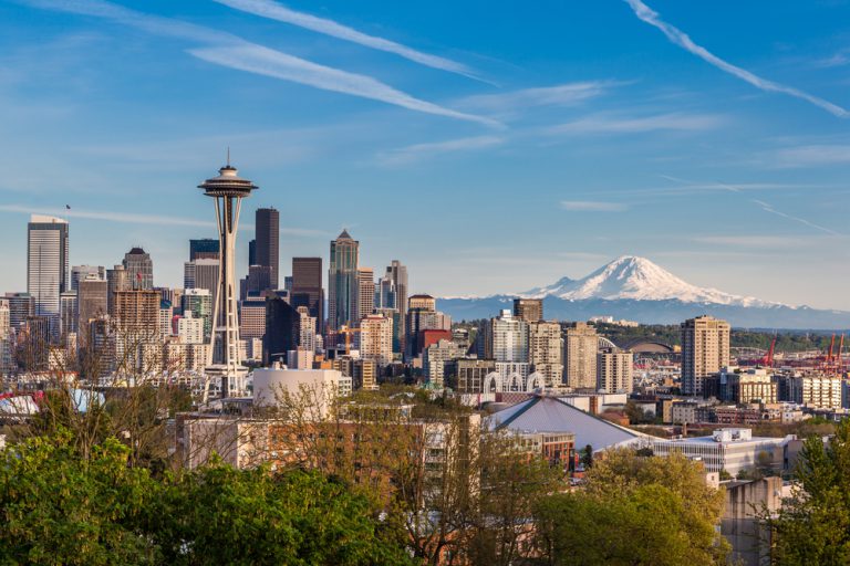 Cityscape with the space needle building and a looming mountain in the distance under a blue sky with wispy white clouds.