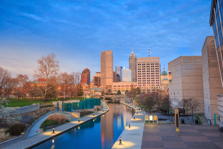 A canal bordered by sidewalks and lights runs through a park and into a cityscape