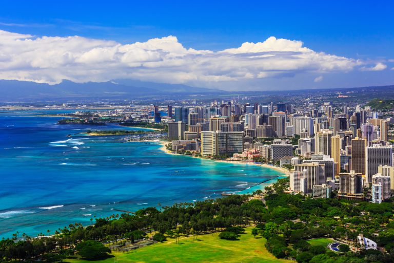 Turquoise blue ocean waters curve along a tree-lined shore that also includes dozens of tall buildings in the background.