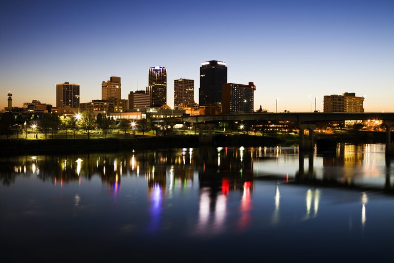 The lights and towers of a city skyline during false dawn that are reflected in calm dark waters