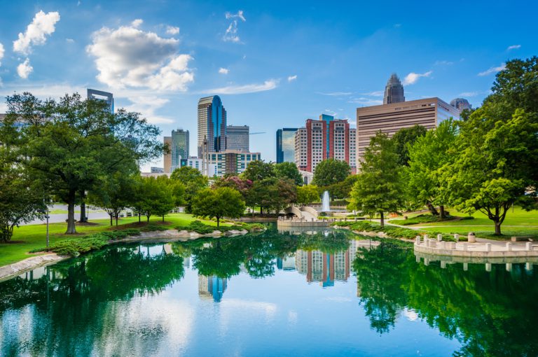 Skyline of a city peaks through the trees surrounding a lake. The sidewalk bordering the lake leads to a water feature.