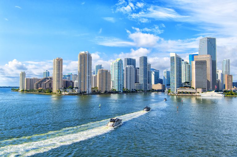 Skyscrapers under a cloudy blue sky standing next to a calm blue ocean with a white boat sailing toward them