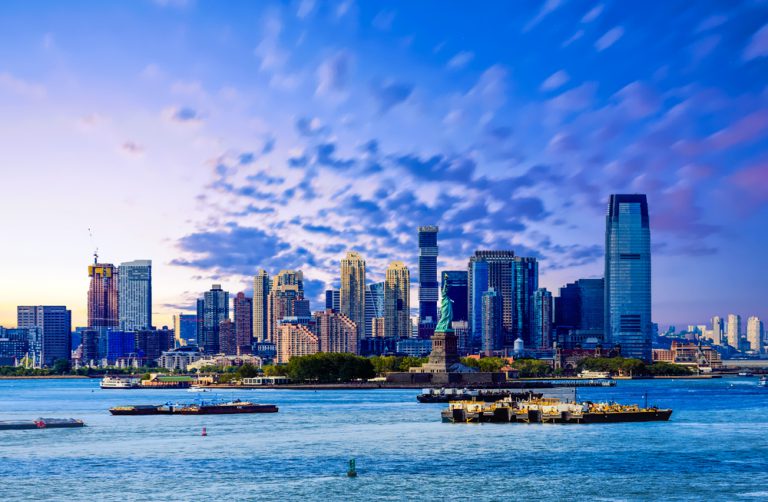 A city skyline under purple and blue sky. Boats float in the Harbor around the Statue of Liberty.