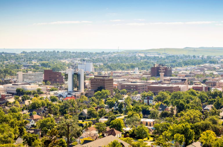 A city with buildings surrounded by green trees under a pale blue sky.