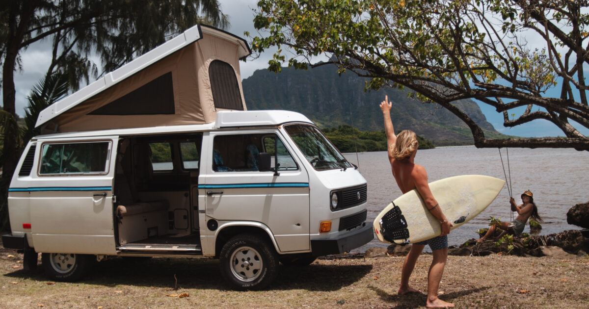 man holding a surfboard next to a campervan