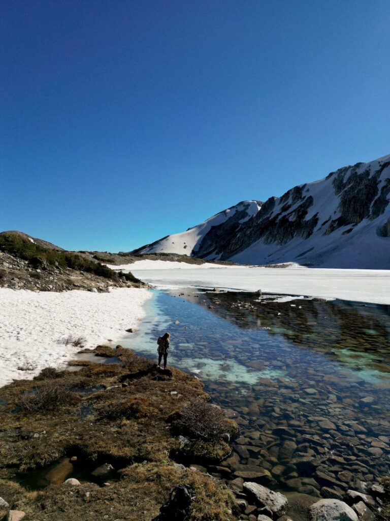 person looking into clear waters surrounded by snowy mountains